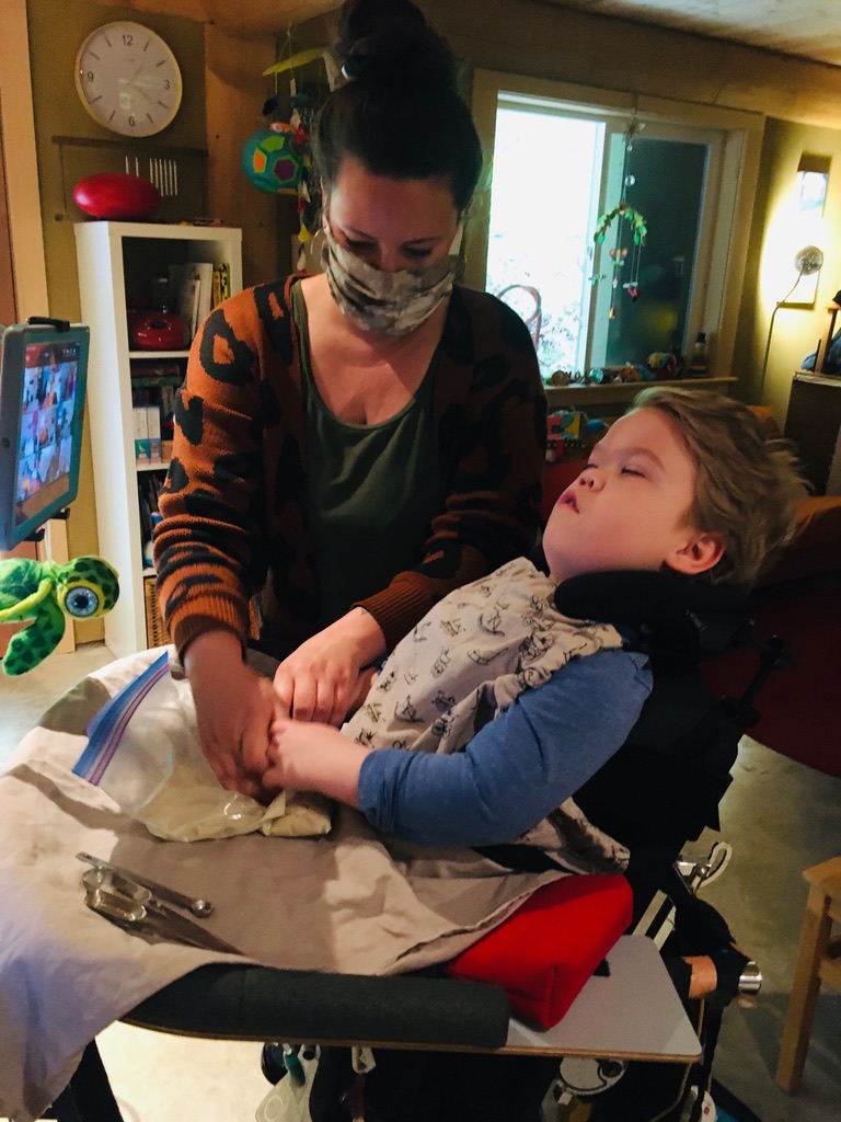 A young boy in a wheelchair is wearing a blue shirt and white bib. He is looking at his computer mounted in front of him. His Intervenor has dark hair tied in a bun and is wearing a mask over her mouth and a green shirt with an orange and black patterned cardigan. The Intervenor is supporting the boy’s hands while they squish white dough in a Ziploc bag.