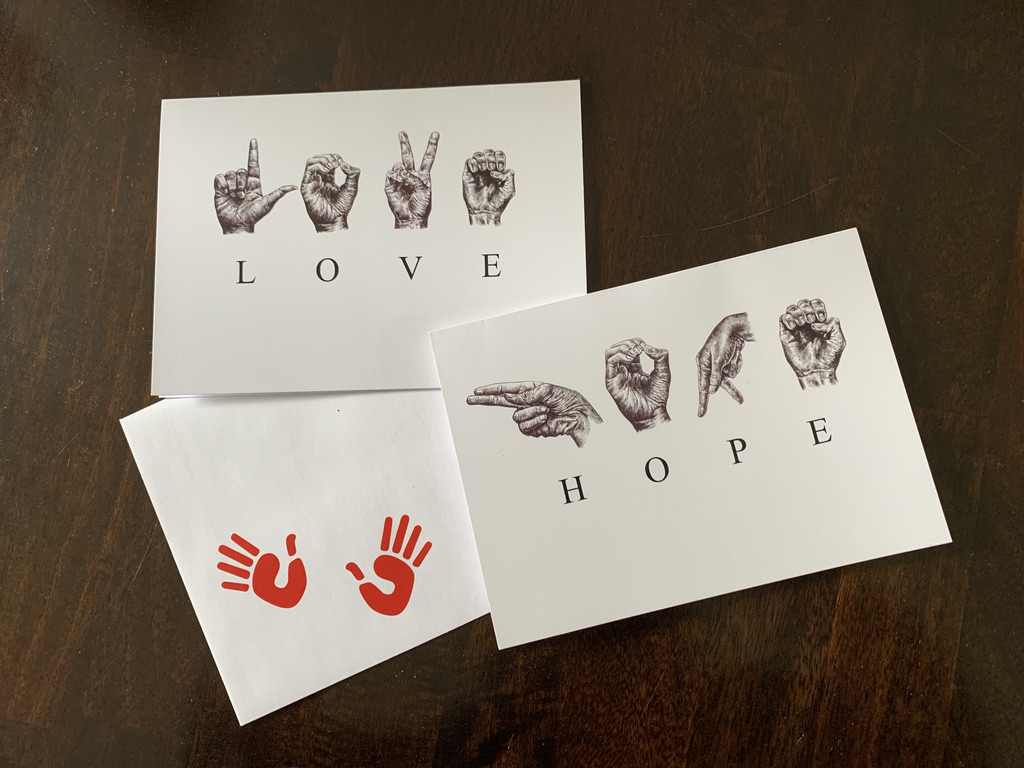 Photo of 2 Greeting cards and 1 envelope with two red hand prints on it. One card has the sketched ASL letters to spell ‘HOPE’ and one card has the sketched ASL letters to spell ‘LOVE’.