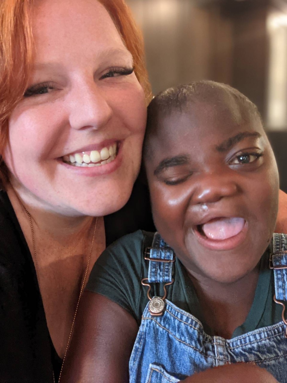 Woman with red hair is smiling brightly. In front of her is a young woman whose head is resting on her cheek with one eye closed wearing a wide smile.